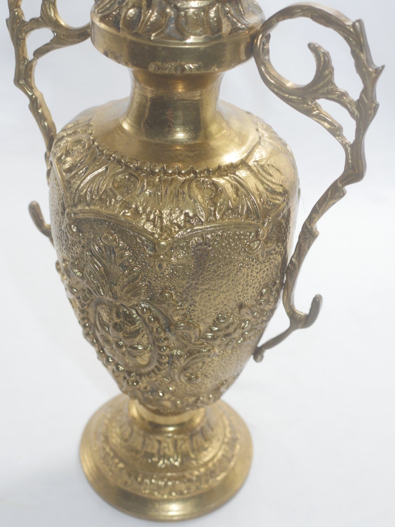 May include: A gold metal vase with intricate floral designs and two ornate handles. The vase has a round base and a tall, slender body.