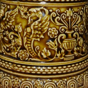 Może przedstawiać: Brązowy kubek ceramiczny z bogatym, tłoczonym wzorem przedstawiającym smoka, kwiaty i wazę. Kubek ma fakturowany brzeg i podstawę.