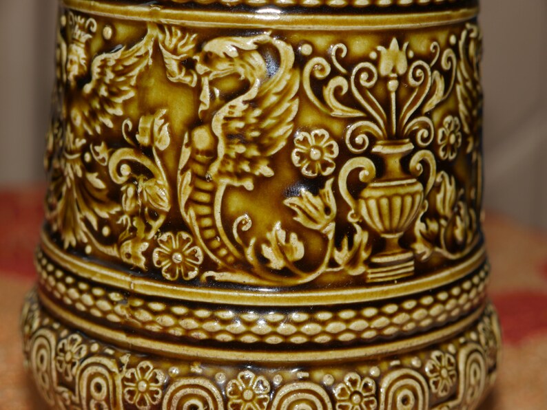 Może przedstawiać: Brązowy kubek ceramiczny z bogatym, tłoczonym wzorem przedstawiającym smoka, kwiaty i wazę. Kubek ma fakturowany brzeg i podstawę.