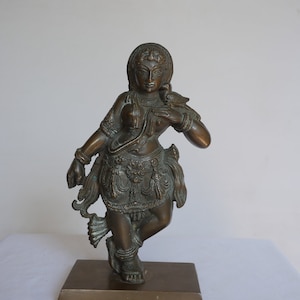 May include: A bronze sculpture of a female figure with intricate details, standing on a rectangular base. The figure is adorned with jewelry and a flowing garment, and holds a bird in her hand.