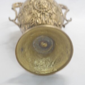 May include: A gold metal vase with an ornate design and two handles. The vase is empty and the bottom is visible.