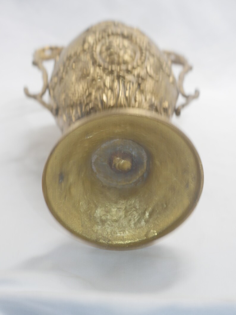 May include: A gold metal vase with an ornate design and two handles. The vase is empty and the bottom is visible.