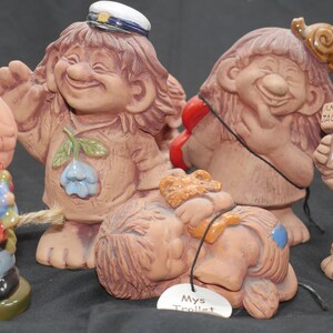 Pu&ograve; includere: Una collezione di cinque figurine di troll in ceramica. I troll sorridono tutti e hanno espressioni diverse. Un troll indossa un cappello da marinaio blu e tiene una corda. Un altro troll tiene un telefono rosso. Un terzo troll tiene un cartello che dice "En Vag Tagard". Un quarto troll sta dormendo e ha un'etichetta che dice "Mys Trollt". Il quinto troll indossa un abito blu e giallo e tiene un mazzo di fiori.