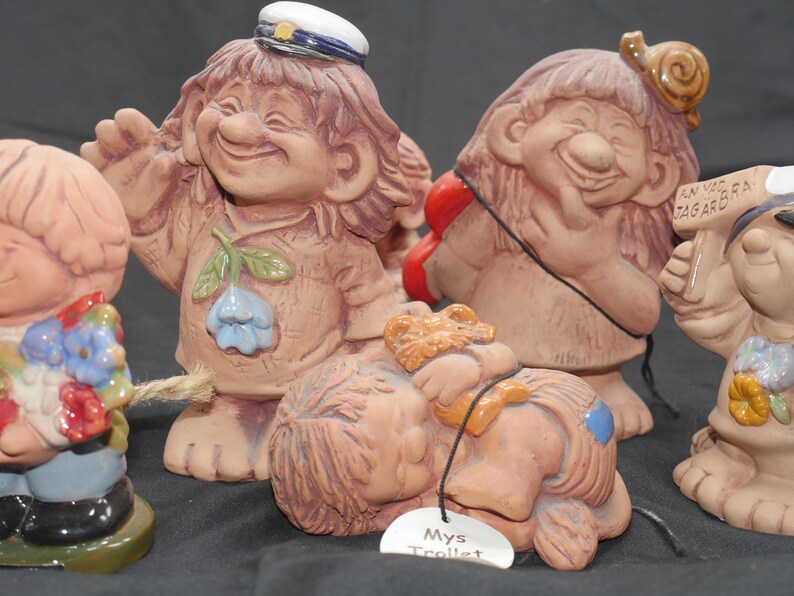 Pu&ograve; includere: Una collezione di cinque figurine di troll in ceramica. I troll sorridono tutti e hanno espressioni diverse. Un troll indossa un cappello da marinaio blu e tiene una corda. Un altro troll tiene un telefono rosso. Un terzo troll tiene un cartello che dice "En Vag Tagard". Un quarto troll sta dormendo e ha un'etichetta che dice "Mys Trollt". Il quinto troll indossa un abito blu e giallo e tiene un mazzo di fiori.