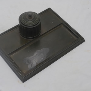 May include: A dark bronze inkwell set. The set includes a rectangular tray with a recessed area and a round inkwell with a lid. The inkwell has a ribbed design. The set is made of metal.
