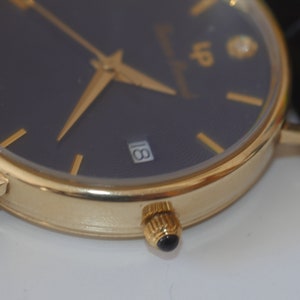 May include: A gold-tone wristwatch with a black face and gold-tone hands. The watch has a date window at the 3 o'clock position and a diamond at the 12 o'clock position.