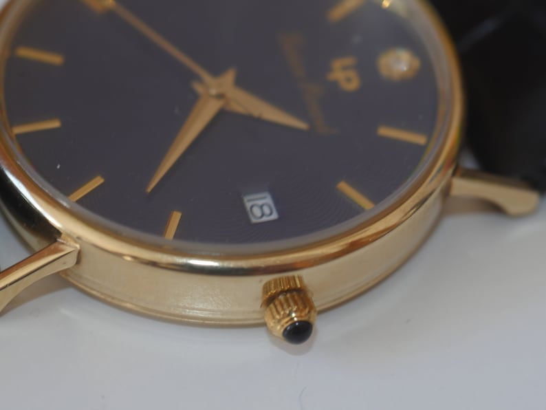 May include: A gold-tone wristwatch with a black face and gold-tone hands. The watch has a date window at the 3 o'clock position and a diamond at the 12 o'clock position.
