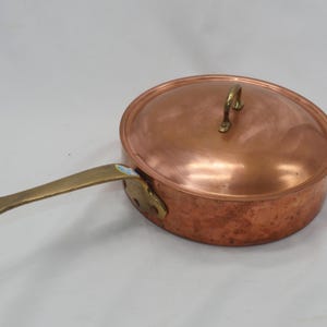 May include: A copper saucepan with a brass handle and lid. The saucepan has a hammered finish and is suitable for cooking on the stovetop.