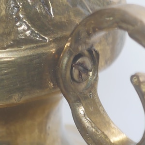 May include: Close-up of a brass hook with a screw. The hook is attached to a decorative object with a textured surface.