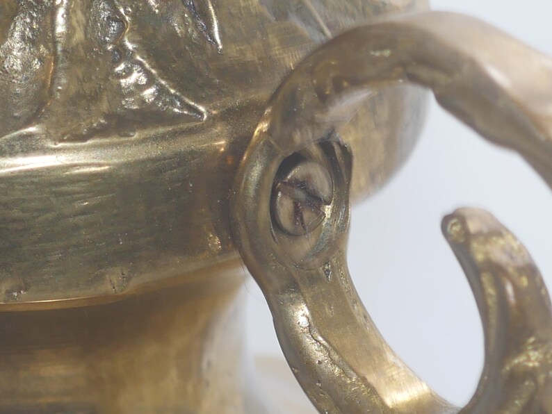 May include: Close-up of a brass hook with a screw. The hook is attached to a decorative object with a textured surface.