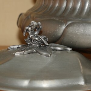 May include: A metallic silver-toned lidded container featuring a sculpted cherub figure perched atop a leaf-shaped finial. The lid has a smooth, rounded surface with a subtle sheen, while the main body displays a ribbed, fluted design.