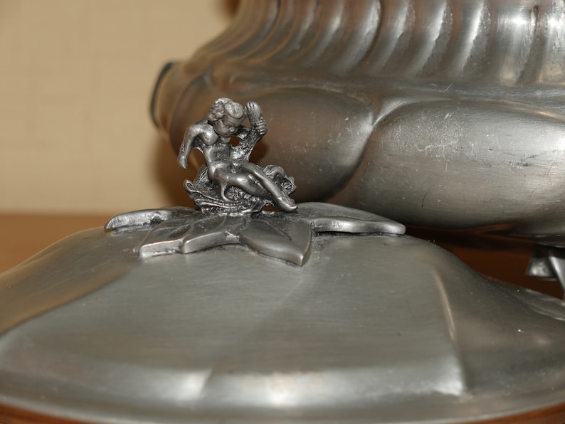 May include: A metallic silver-toned lidded container featuring a sculpted cherub figure perched atop a leaf-shaped finial. The lid has a smooth, rounded surface with a subtle sheen, while the main body displays a ribbed, fluted design.