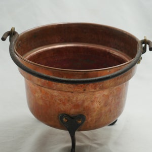 May include: A copper pot with a black iron handle and three legs. The pot has a hammered finish and is in good condition.