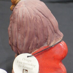 Pu&ograve; includere: Una statuetta in ceramica di una donna con lunghi capelli castani che indossa un'ala rossa a forma di cuore. La statuetta ha una lumaca sulla testa e un'etichetta bianca che recita "Design Ewa Jarenskog Jie-Keramik".