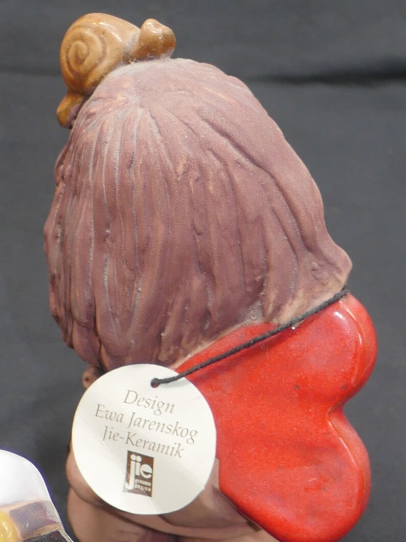 Pu&ograve; includere: Una statuetta in ceramica di una donna con lunghi capelli castani che indossa un'ala rossa a forma di cuore. La statuetta ha una lumaca sulla testa e un'etichetta bianca che recita "Design Ewa Jarenskog Jie-Keramik".
