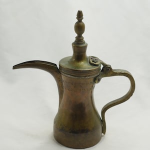 Op de afbeelding: Een koperen koffiepot met een scharnierend deksel en een decoratieve handgreep. De pot heeft een traditioneel Arabisch ontwerp en is in goede staat.