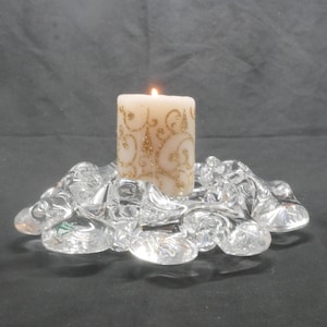 Peut inclure: Une bougie pilier ivoire allumée avec des tourbillons de paillettes dorées repose sur un bougeoir en verre transparent. Le bougeoir a une forme organique. Le fond est gris foncé.