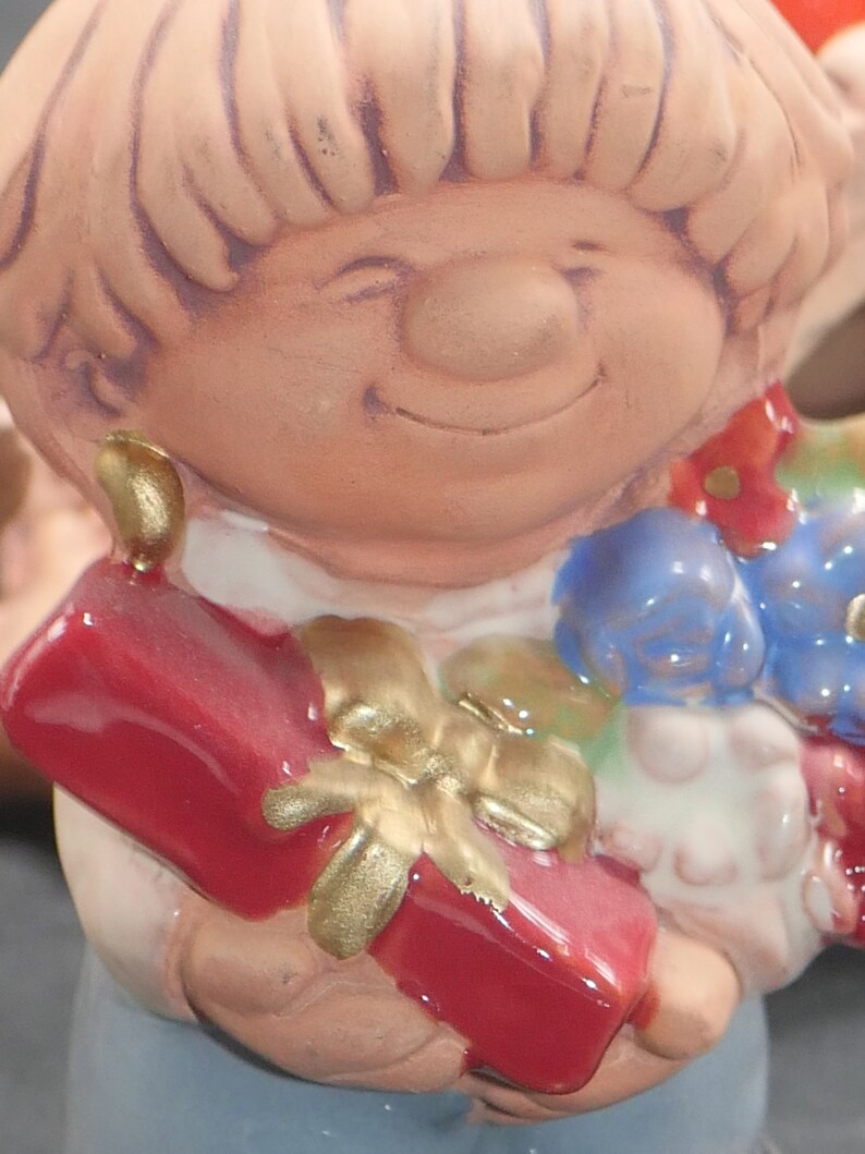 Pu&ograve; includere: Una statuetta in ceramica di una ragazza sorridente con i capelli biondi che tiene una scatola regalo rossa con un fiocco d'oro. La ragazza indossa un vestito bianco con fiori blu.