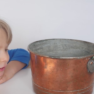 May include: A young child looks intently at a large copper pot with a handle. The pot has a shiny, aged copper finish and a silver lining inside.