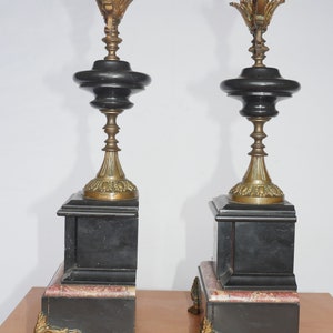 May include: A pair of black and gold decorative fireplace mantle clocks with ornate details. Each clock has a black marble base with a gold-colored top and a pink marble accent.