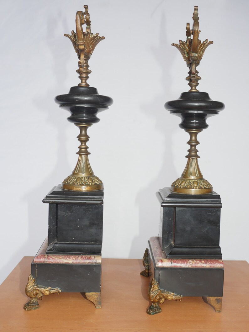 May include: A pair of black and gold decorative fireplace mantle clocks with ornate details. Each clock has a black marble base with a gold-colored top and a pink marble accent.
