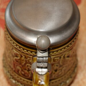 Może przedstawiać: Brązowy ceramiczny kufel do piwa z pokrywką z cyny i zawiasowym uchwytem. Kufel jest ozdobiony wzorem kwiatowym.