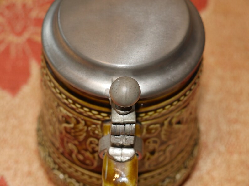 Może przedstawiać: Brązowy ceramiczny kufel do piwa z pokrywką z cyny i zawiasowym uchwytem. Kufel jest ozdobiony wzorem kwiatowym.