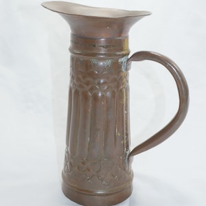 Könnte beinhalten: Eine Kupferkanne mit einem verzierten Design. Die Kanne hat eine breite Öffnung und einen Henkel. Die Kanne ist aus gehämmertem Kupfer und hat eine braune Patina.