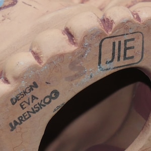 Pu&ograve; includere: Primo piano della parte inferiore di una statuetta in ceramica color terracotta. La statuetta ha un foro sul fondo e il testo "DESIGN EVA JARENSKOO" e "JIE" sono stampati sul fondo.