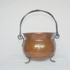 May include: A copper cauldron with a hammered texture, featuring a dark metal handle and three small feet. The cauldron has a rounded body and a shiny, reflective surface, perfect for decorative use.