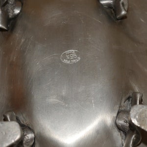May include: A silver-toned metal bowl with a smooth, brushed surface and four sturdy, textured feet. An oval hallmark is stamped on the underside, featuring the text "ETAIN FIN 95% ZINN" in a circular arrangement, indicating the metal composition.