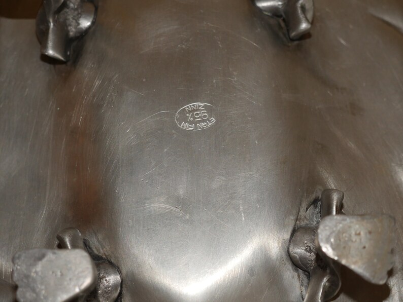May include: A silver-toned metal bowl with a smooth, brushed surface and four sturdy, textured feet. An oval hallmark is stamped on the underside, featuring the text "ETAIN FIN 95% ZINN" in a circular arrangement, indicating the metal composition.