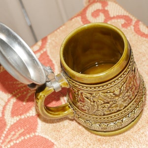 Może przedstawiać: Zielony ceramiczny kufel do piwa z pokrywką z cyny. Kufel ma szczeg&oacute;łowy wz&oacute;r kwiatowy i uchwyt.