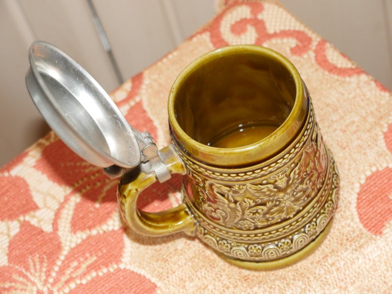 Może przedstawiać: Zielony ceramiczny kufel do piwa z pokrywką z cyny. Kufel ma szczeg&oacute;łowy wz&oacute;r kwiatowy i uchwyt.