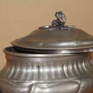May include: A metallic gray pewter tureen with a fluted body and a matching lid. The lid features a sculptural finial depicting a figure riding a dolphin. The vessel has a wide, rounded bowl shape with integrated side handles and a smooth finish.