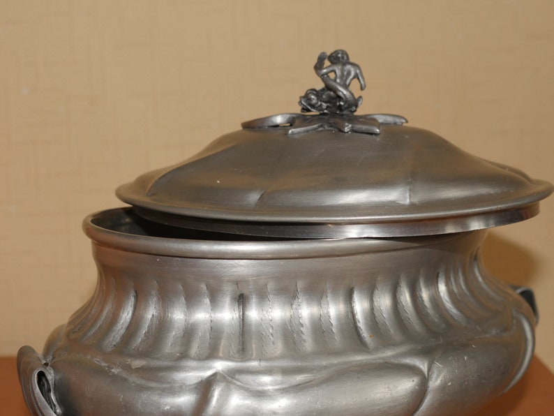 May include: A metallic gray pewter tureen with a fluted body and a matching lid. The lid features a sculptural finial depicting a figure riding a dolphin. The vessel has a wide, rounded bowl shape with integrated side handles and a smooth finish.