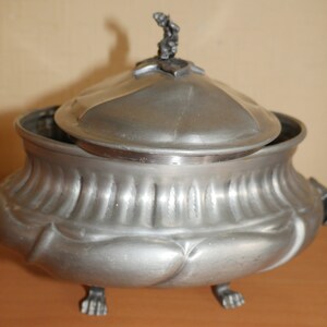 May include: A metallic gray lidded tureen with a matte finish, featuring a rounded body with vertical fluted detailing on the upper section and smooth, bulbous lobes on the lower portion. It stands on three claw-style feet and has a small finial.