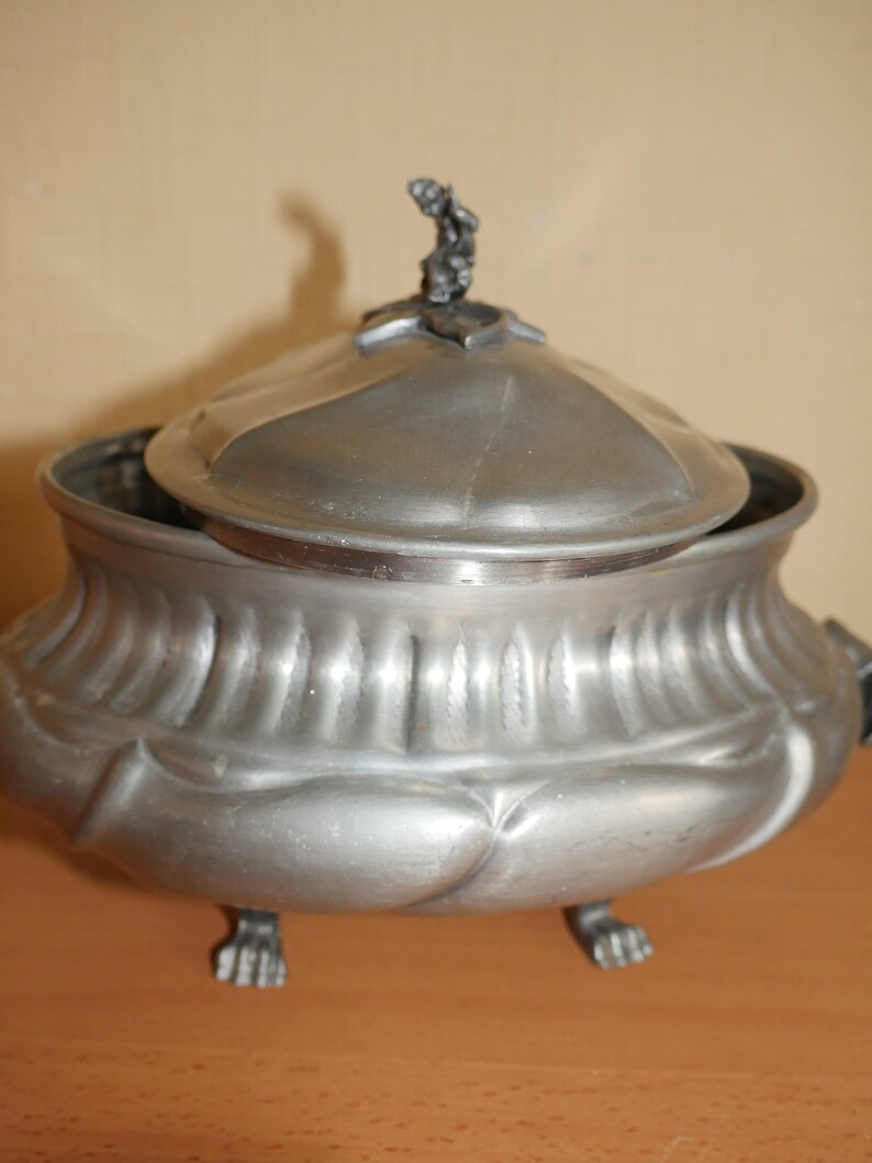 May include: A metallic gray lidded tureen with a matte finish, featuring a rounded body with vertical fluted detailing on the upper section and smooth, bulbous lobes on the lower portion. It stands on three claw-style feet and has a small finial.