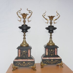 May include: A pair of black and gold decorative candle holders with ornate details. Each holder features a black marble base with gold accents and a gold metal candle holder with a floral design.