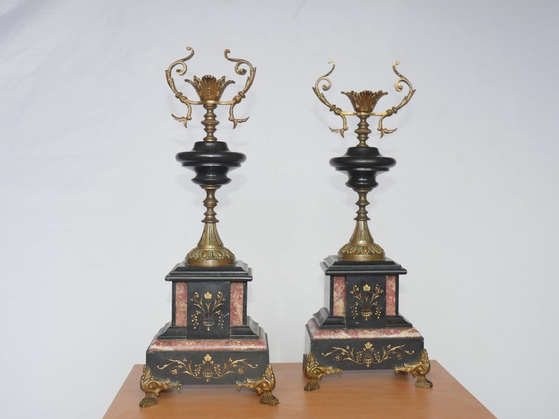 May include: A pair of black and gold decorative candle holders with ornate details. Each holder features a black marble base with gold accents and a gold metal candle holder with a floral design.