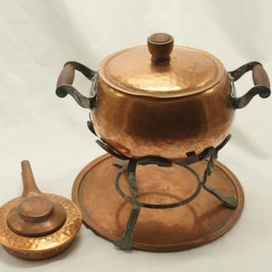 Può includere: Una pentola per fonduta in rame con coperchio e dettagli con manico in legno. La pentola poggia su un supporto in rame abbinato con una base circolare. Una piccola padella in rame con manico è accanto alla pentola per fonduta. Il set è progettato per cucinare e servire.