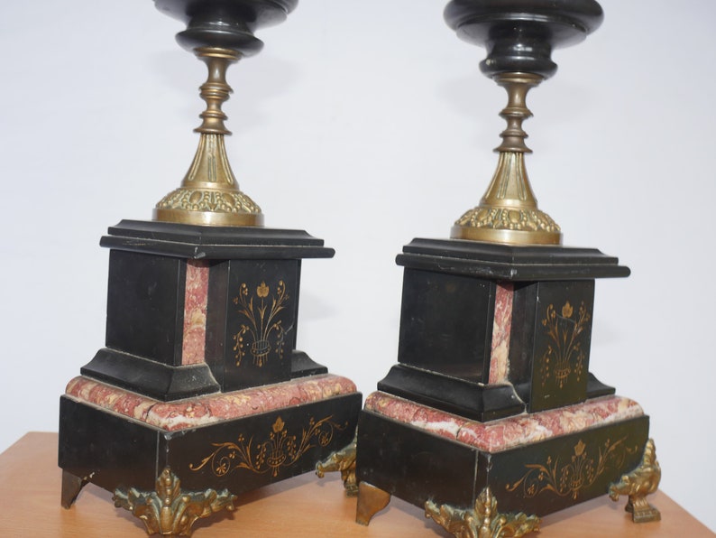 May include: A pair of black and gold candle holders with marble bases. The bases are decorated with gold floral designs.