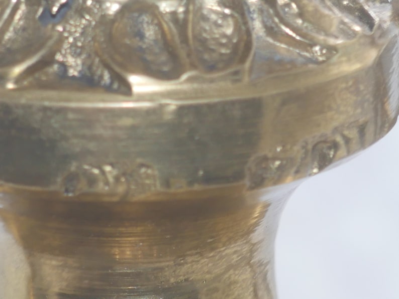 May include: Close-up of the bottom of a brass candlestick with an ornate design. The bottom of the candlestick has a small inscription that reads "Made in Italy".