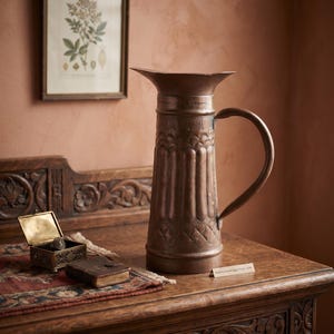 May include: A tall copper pitcher with a flared rim and a curved handle, featuring embossed vertical fluting and geometric patterns around the body. The pitcher sits on a wooden surface next to a small open metal box and a miniature leather-bound book.