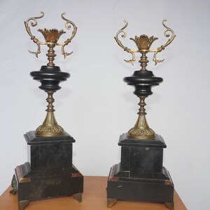 May include: A pair of black and gold decorative candle holders with ornate details. The candle holders are made of metal and have a black marble base.