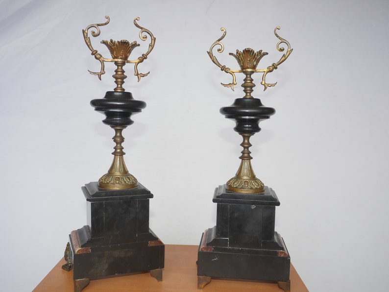 May include: A pair of black and gold decorative candle holders with ornate details. The candle holders are made of metal and have a black marble base.