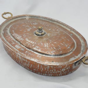 May include: An oval copper serving dish with a lid and two brass handles. The dish has a hammered finish and a tarnished patina.