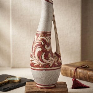 Peut inclure: Un grand vase en céramique avec une silhouette incurvée et une anse intégrée. La surface présente une base blanc cassé mouchetée avec une bande centrale ornée de motifs à volutes en relief rouge foncé et dorés.