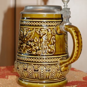 Może przedstawiać: Brązowy ceramiczny kufel do piwa z pokrywką i uchwytem z cyny. Kufel jest ozdobiony wzorem kwiatowym i parą skrzydlatych stworzeń.