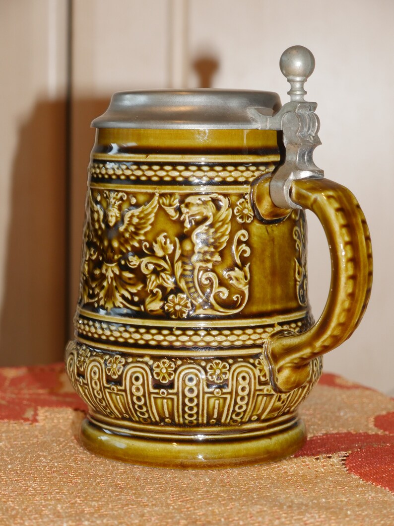Może przedstawiać: Brązowy ceramiczny kufel do piwa z pokrywką i uchwytem z cyny. Kufel jest ozdobiony wzorem kwiatowym i parą skrzydlatych stworzeń.